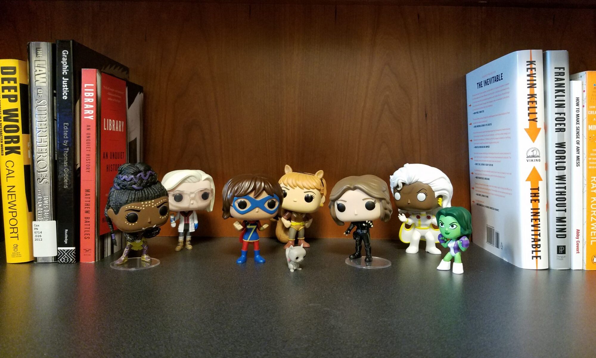 Funko Pop figures of women superheroes lined up on a bookshelf between books on libraries, law, and technology